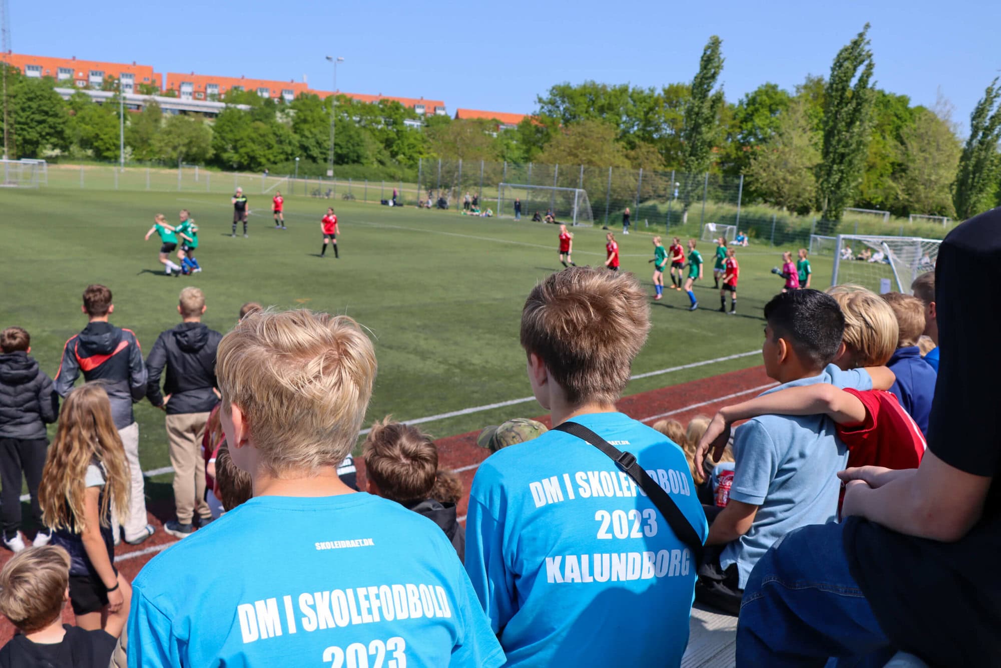 Børn på en tribune sidder og ser en fodboldkamp til DM i skolefodbold 2023